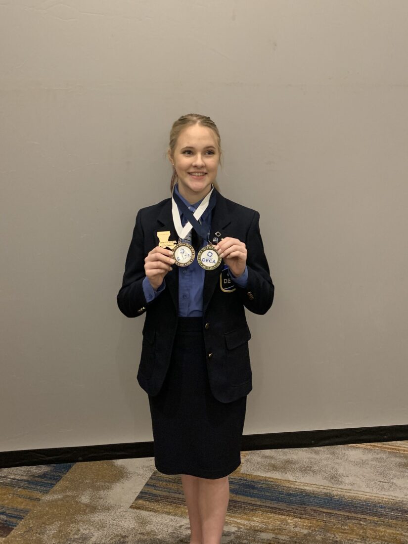 A person in a blue blazer and skirt stands on a patterned carpet, holding two medals to celebrate online school success.