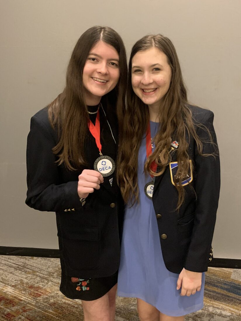 Two young women in blazers and DECA medals smile, celebrating their online charter school in front of a plain wall.