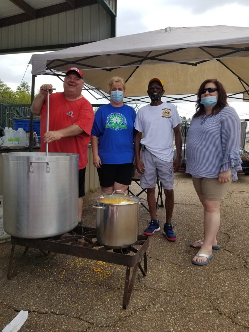 Four adults cook together under a canopy outdoors, talking near two large pots on propane burners.