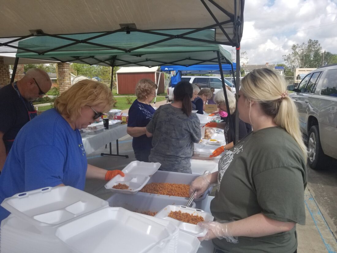 Volunteers wearing gloves assemble rice meal boxes under tents at an outdoor K-12 online charter school event.