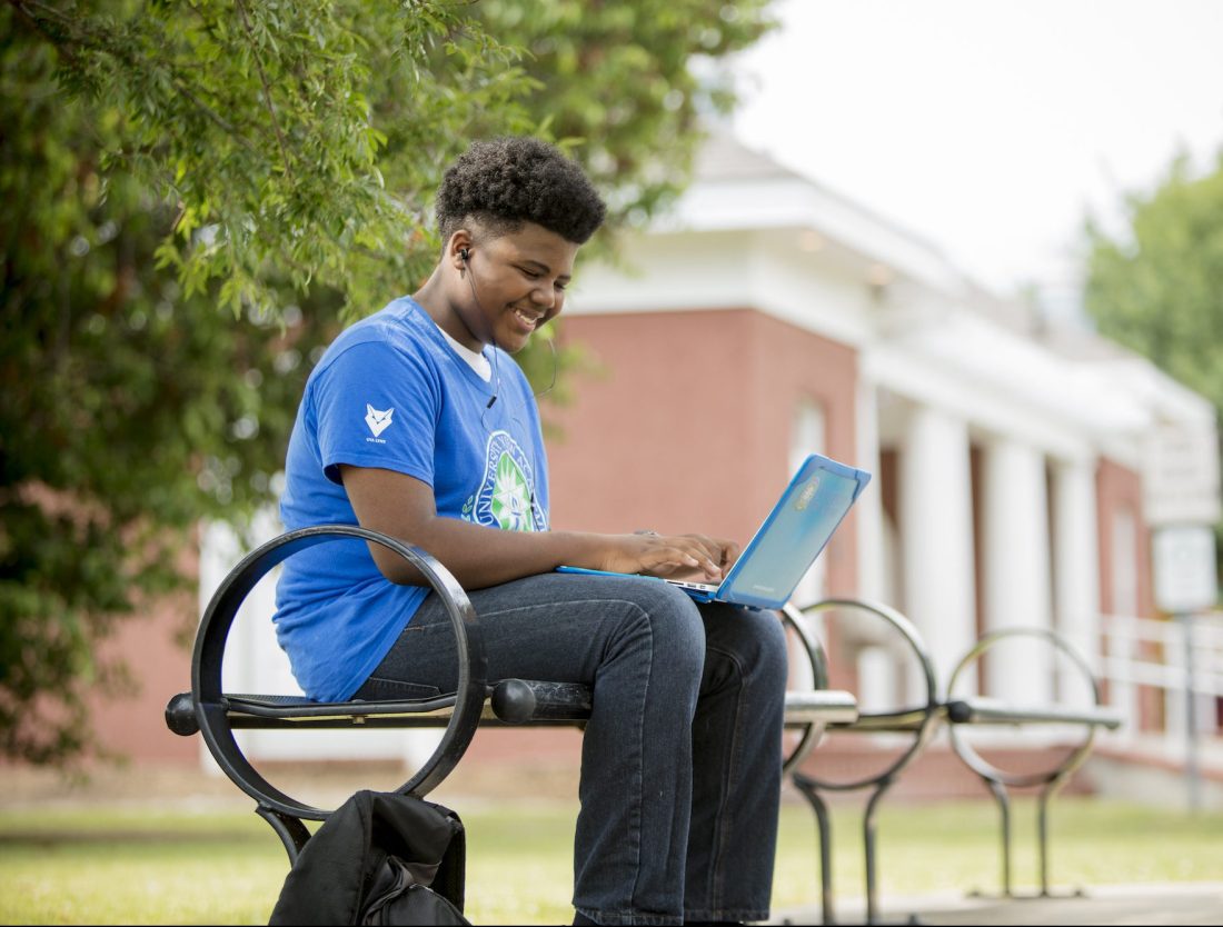 A teenage boy seated on a bench, working on a laptop.