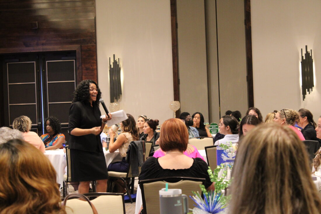 A woman speaking in front of a crowded event hall.