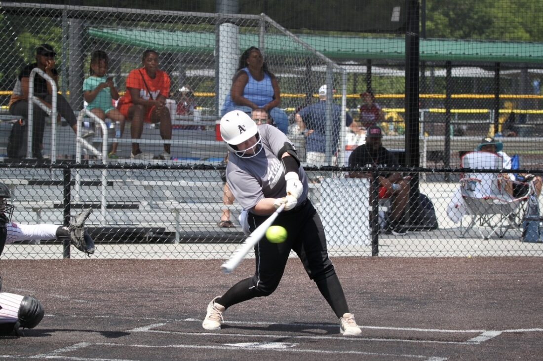 An action photograph of a girl in softball gear swinging a bat at the ball.