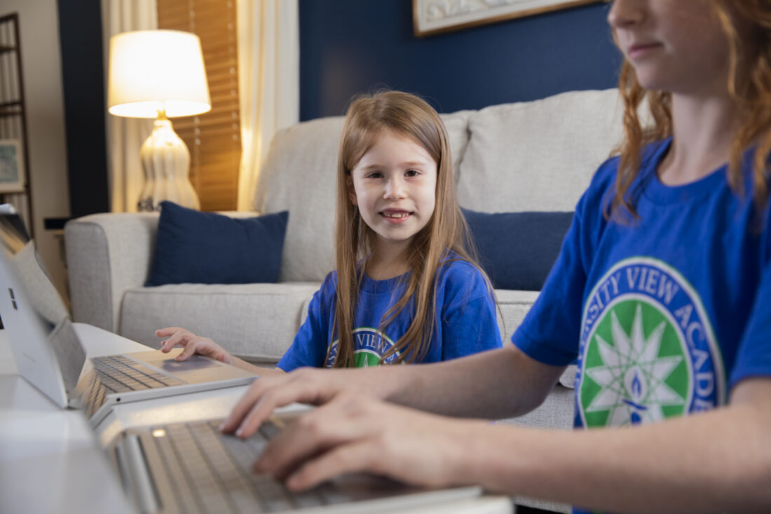 Two kids in matching blue shirts use laptops for K-12 online classes at a table in a cozy living room.