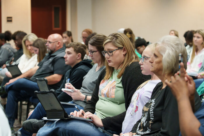 A parent takes notes on a laptop at orientation. She is seated in a row full of attendees.