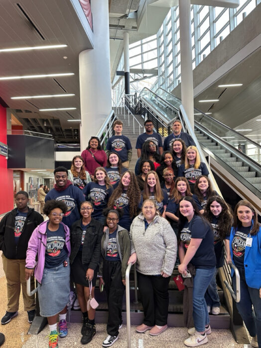 A large group photograph of UVA DECA members and advisors standing on a staircase at the Sports Marketing Conference. They are all wearing Pelicans t-shirts.