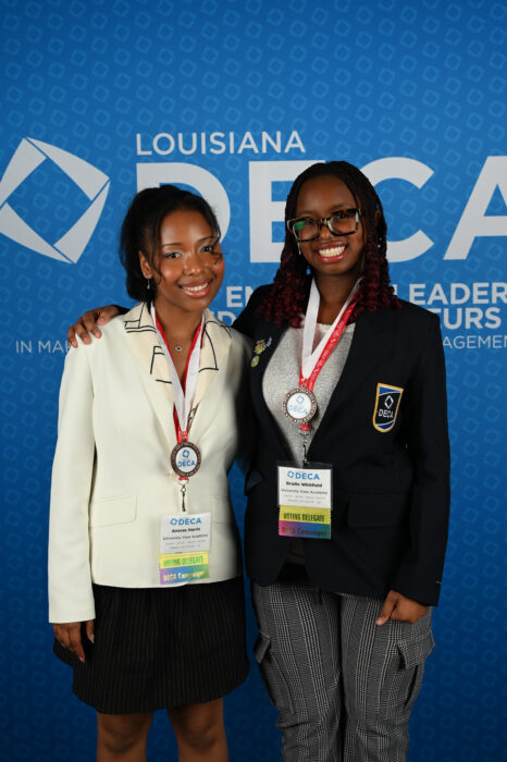 Two female high school DECA members smile for a picture against a blue backdrop with the words Louisiana DECA. They are both sharply dressed with award medallions.
