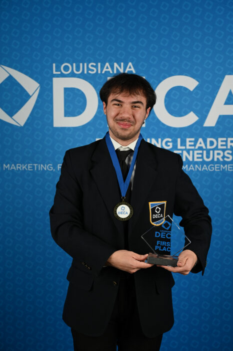 Brock Sykes holds his 1st place award trophy and poses against a blue backdrop with the words Louisiana DECA.