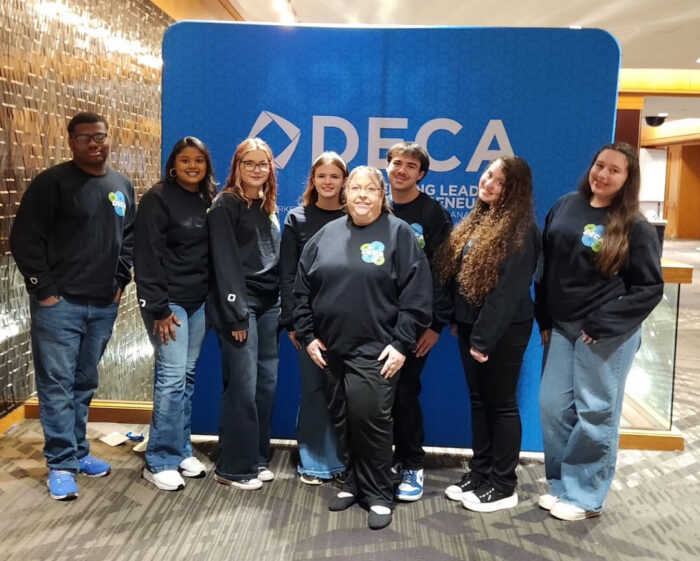 Chapter officers in logoed sweatshirts pose for a group photo with advisor Mindy Rose in front of a blue DECA backdrop.
