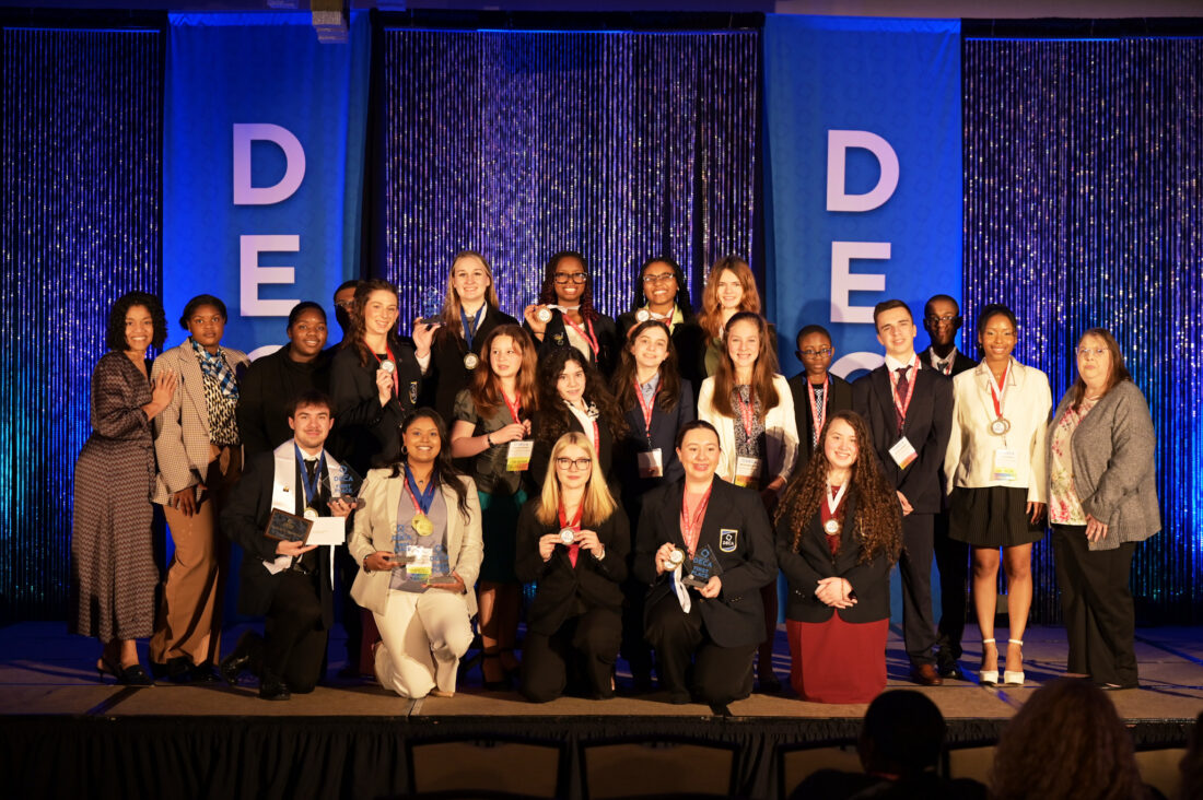 Group photograph of UVA's DECA chapter members on a stage with advisor Mindy Rose and Superintendent Dr. Quentina Timoll. All the students are wearing award medallions and some in the front row are holding trophies.