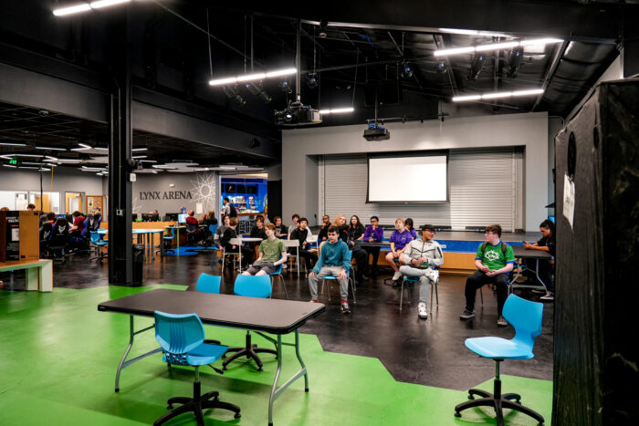 Students in a modern classroom get ready for K-12 online lessons, facing a blank projector screen on green and black floors.