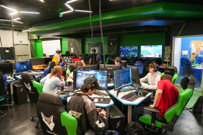 People sit at desks with computers in a gaming room, wearing headsets like K-12 online charter school students.