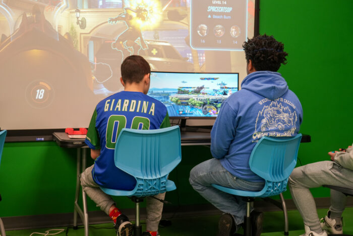 Two people in blue chairs play a video game, illustrating interactive online charter school classes by a green wall.