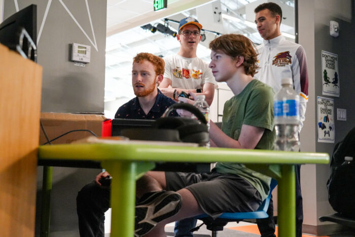 Two people play video games at a green table while others watch, taking a break from online school in a bright room.