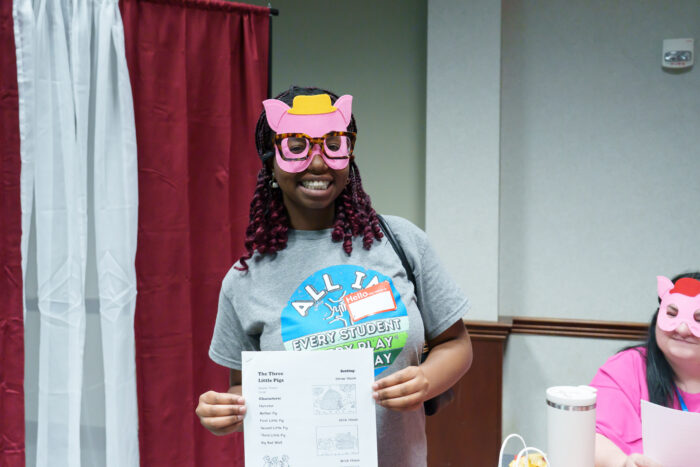 A student holds up the script for The Three Little Pigs while smiling and wearing a felt pig mask