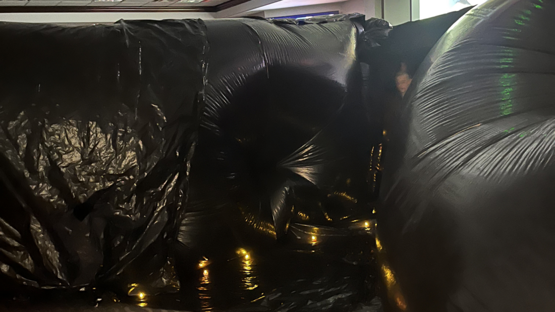 Large black plastic sheets create a tunnel lined with string lights on the floor, in a room with white walls and ceiling lights.
