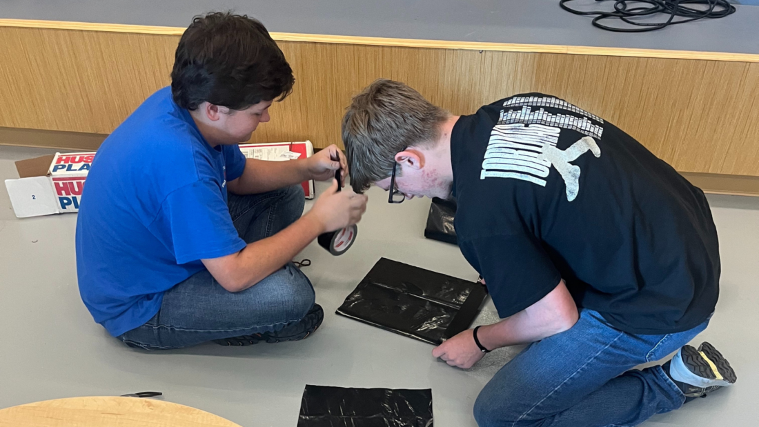 Two boys kneel on the floor, working with black plastic sheets and tape.