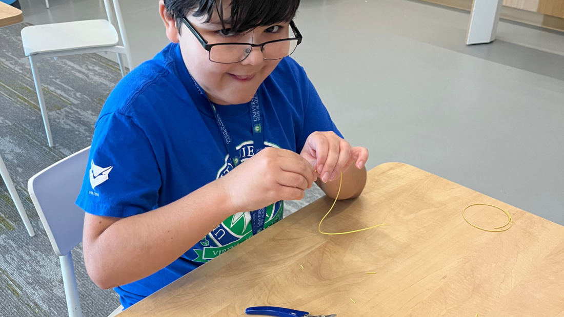 A boy wearing glasses threads yellow wire at a table, with pliers and wire pieces spread out in front of him.