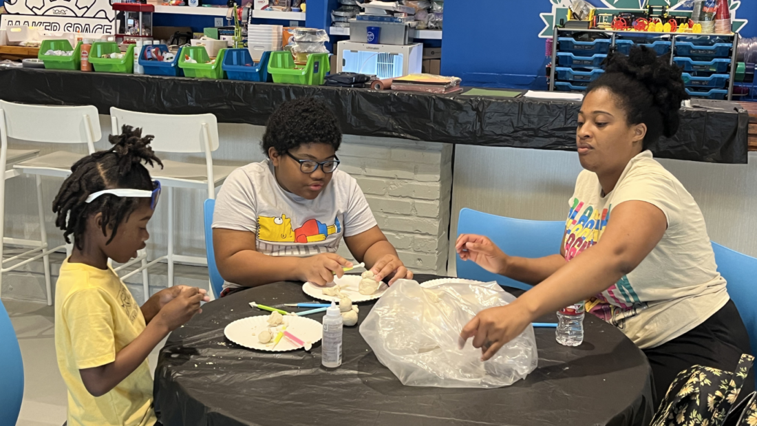 Three people at a round table work on a craft project with white modeling material and tools in a colorful room.