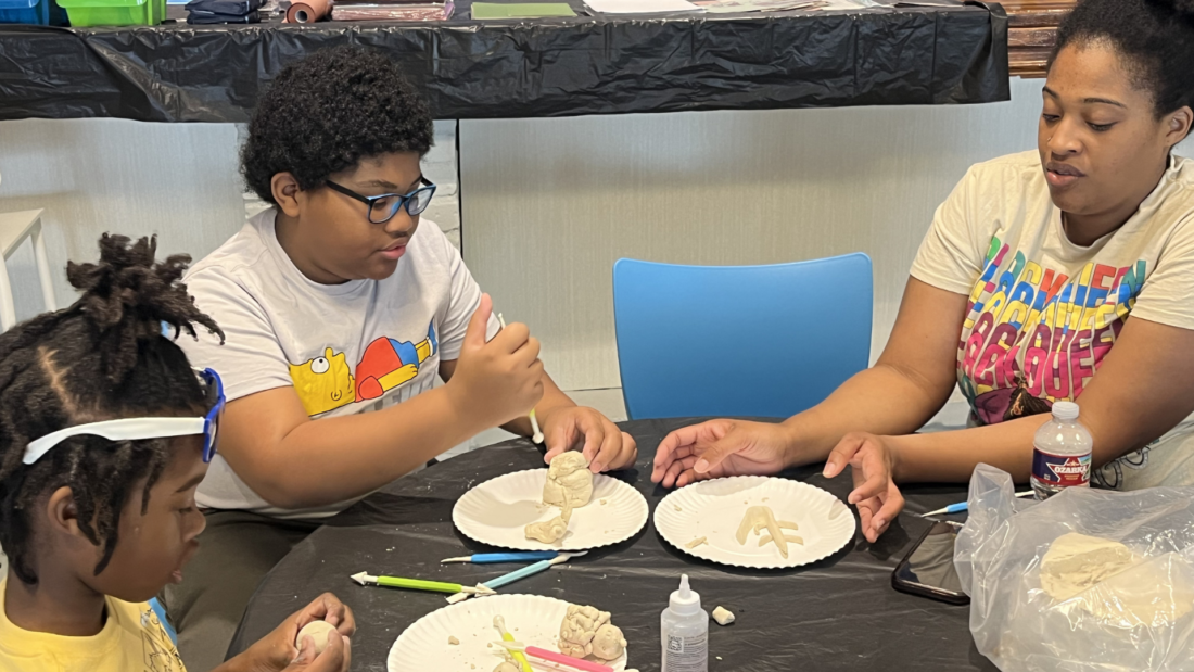 Three people make clay sculptures on paper plates at a table covered with scattered art supplies and tools.