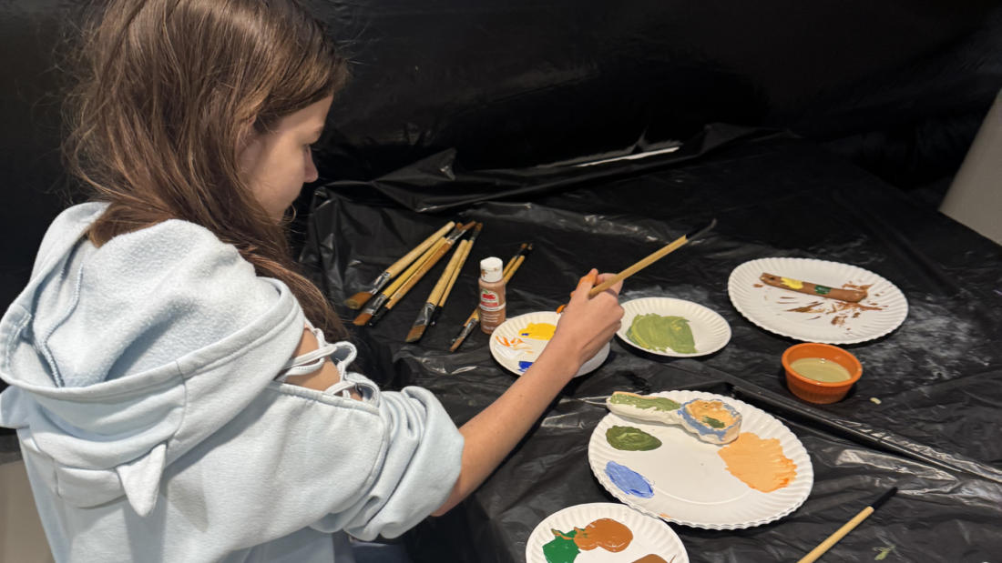 A girl paints ceramics at a table covered with paintbrushes, bottles of paint, and paint-mixed paper plates.