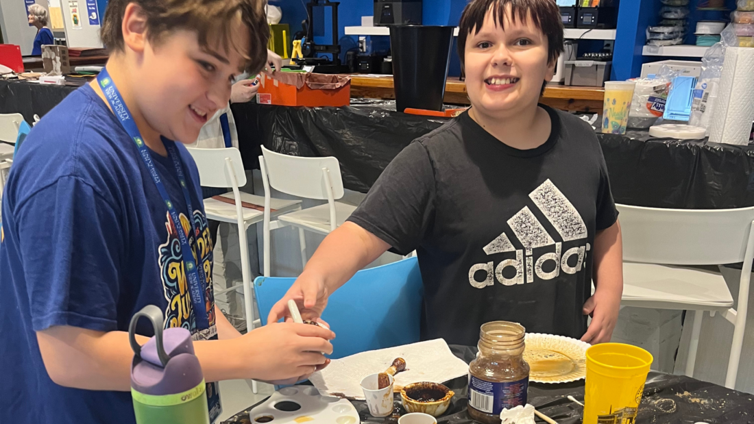 Two smiling kids stand at a table preparing food, surrounded by snacks, dips, jars, cups, and a water bottle.