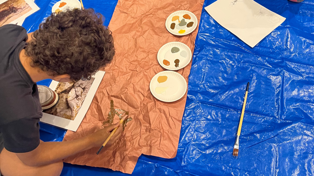 A student uses a brush to paint leaf designs on brown paper, with art supplies and paint palettes on a blue tarp nearby.