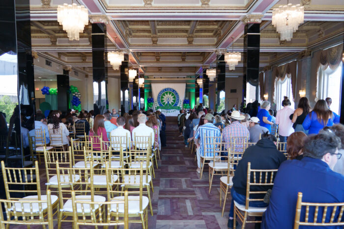 The view from the rear of the room at ring ceremony, many people seated in chairs and looking towards the ornate stage