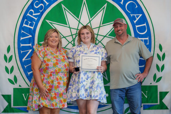 A student holds up a certificate and smiles for a picture with her parents