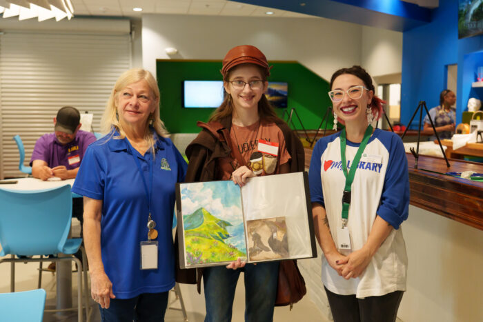 A student holds up her artwork and smiles for a picture with two teachers.