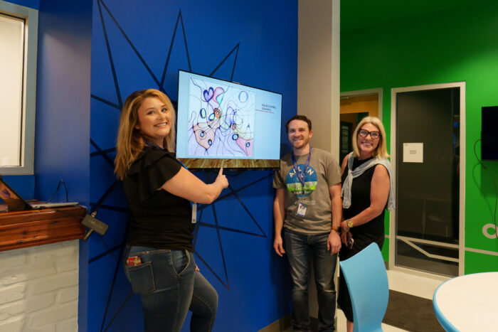 Three teachers smile and point to a student's artwork displayed on a screen.