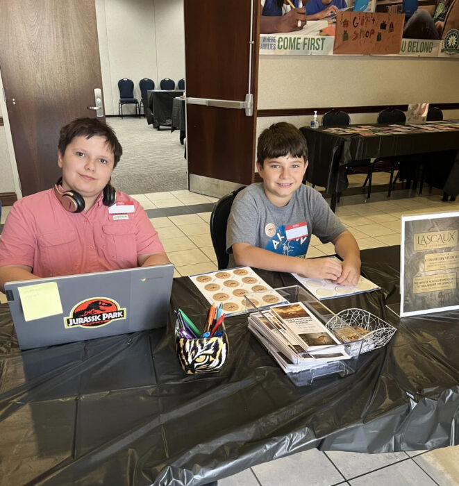 Two students sit behind a table at the museum exhibit, with various signs and items displayed in front of them.