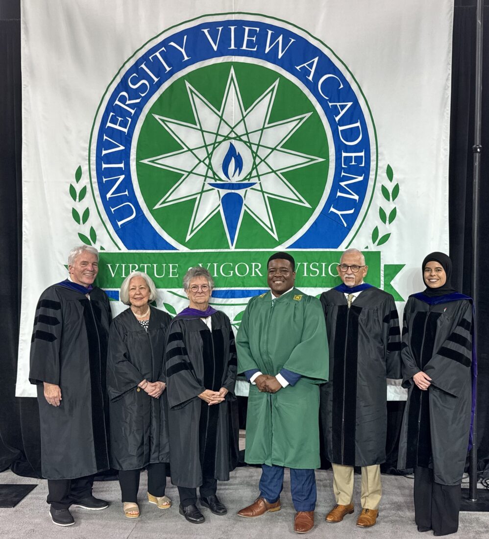 Six people in academic regalia stand beneath a University View Academy banner at the 2025 graduation ceremony.