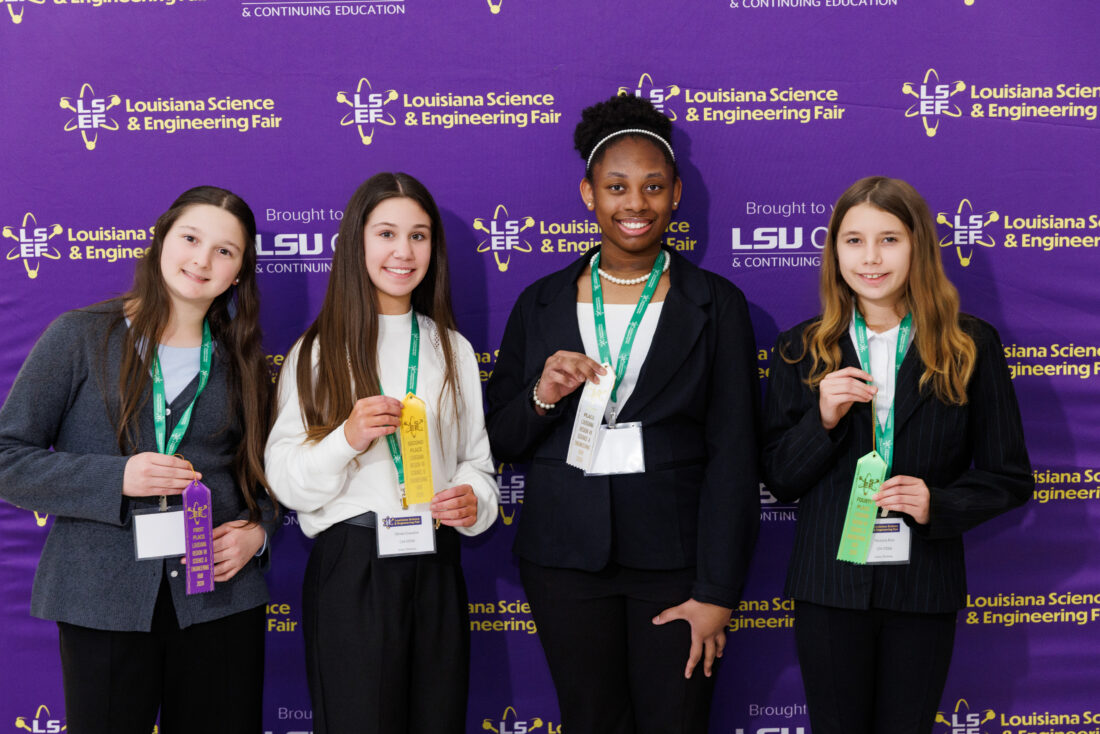 Four girls, including a UVA 8th grader, stand in front of an LSU Science Fair backdrop holding ribbons and badges.