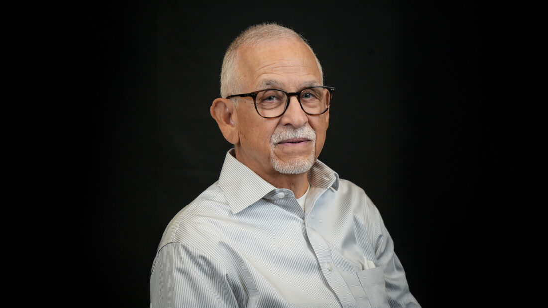 Older man with short gray hair, glasses, and a trimmed beard in a light dress shirt sits before a black background.
