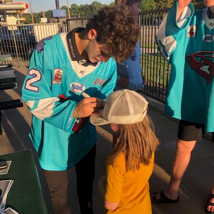 A hockey player in a teal jersey signs a white cap for a young girl in yellow outdoors, chatting with her.