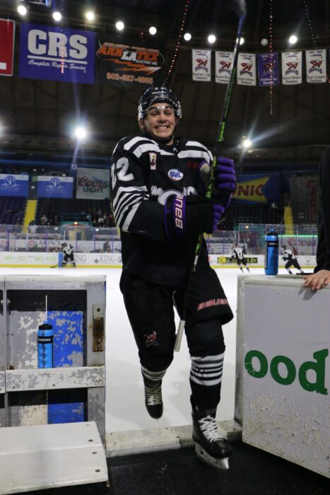 A hockey player in a black uniform smiles as he steps onto the ice rink after finishing his online school classes.