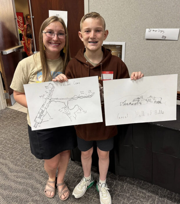 A smiling student holds up his art with a friend
