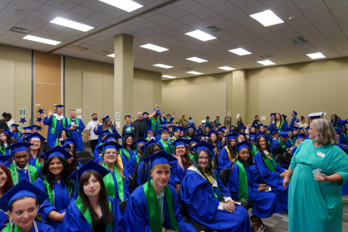 A large group of students in blue and green graduation gowns smile at the camera in a room.
