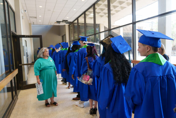 Graduates in blue gowns line up in a hallway to proceed to the arena, guided by a teacher.