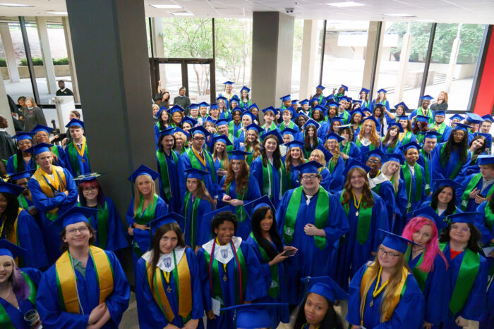 A large group of graduates in blue gowns gather indoors, smiling and looking up at the camera.