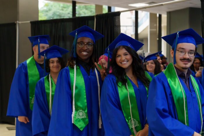 Graduates in blue gowns and green stoles line up indoors, all smiling at the camera.