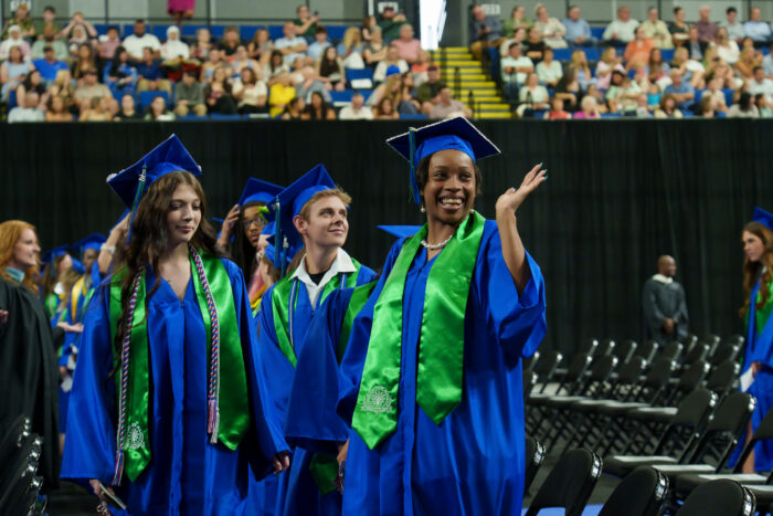 Graduates in blue gowns and green stoles walk to their seats; a student in front waves to the audience.