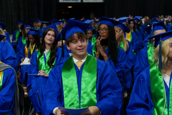 Graduates in blue caps and gowns with green stoles sit, smiling and holding programs during the ceremony.