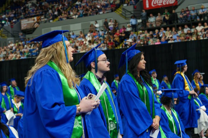 Three graduates in blue caps and gowns stand in a row to be recognized at a crowded ceremony.
