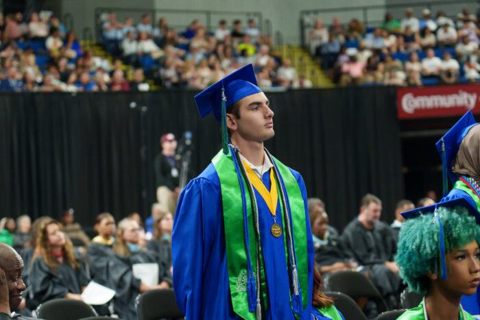 A graduate in a blue cap and gown stands among seated peers to be recognized.