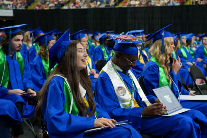 Graduates in blue caps and gowns sit in rows, with one student in front laughing in joy.