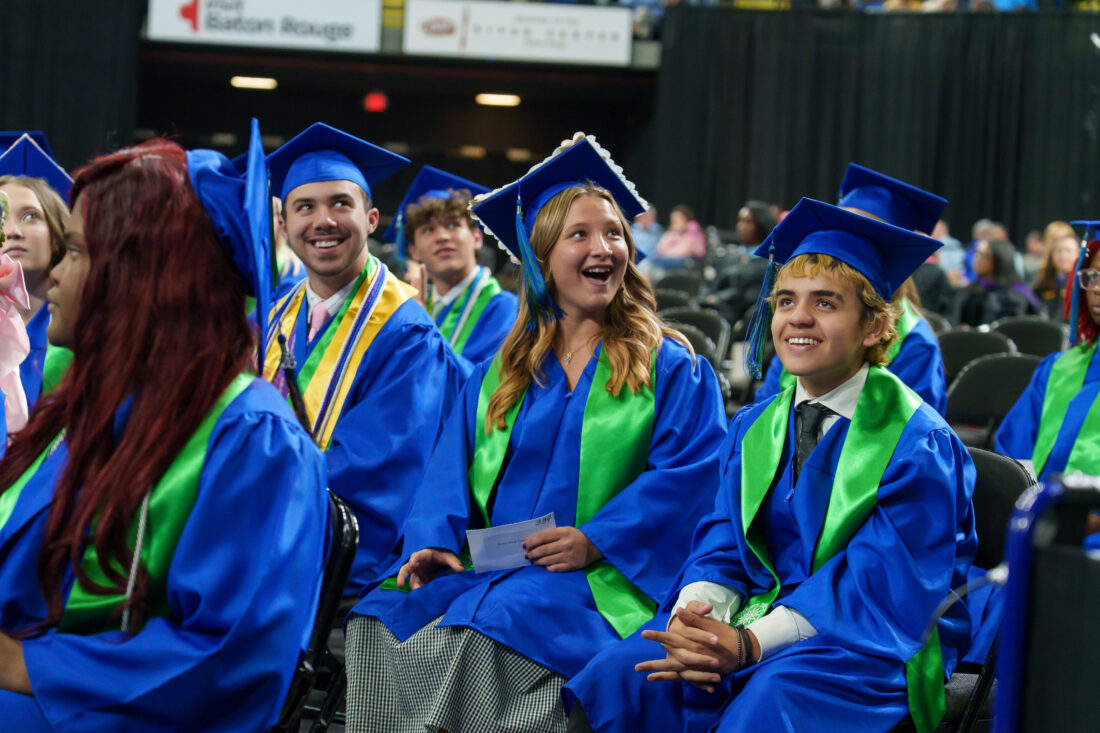 Graduates in blue caps and gowns with green stoles sit at graduation, with one student smiling at the audience.