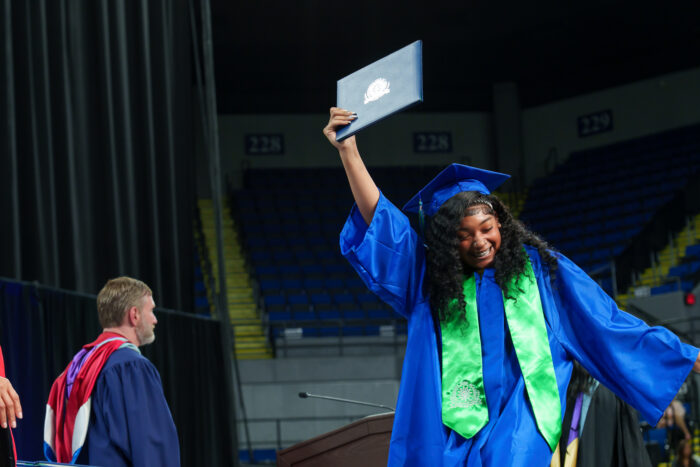 A graduate in a blue cap and gown smiles and holds up her diploma triumphantly.