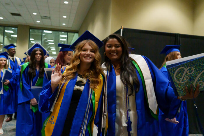Two graduates in blue caps and gowns smile and pose for a picture, diplomas in hand, during the recessional.